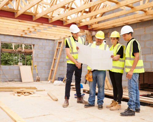 Builder On Building Site Looking At Plans With Apprentices Builder On Building Site Looking At Plans With Apprentices